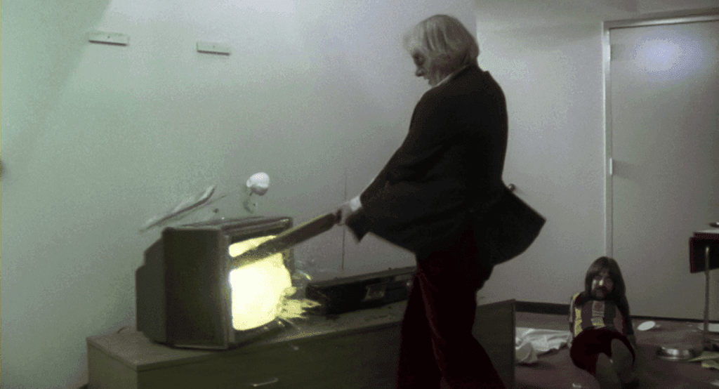 Ian, Spinal Tap's manager, smashing a TV so that it explodes, while Derek Smalls watches.